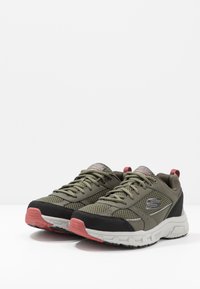 Zapatillas deportivas de malla verde con acentos negros y suelas de goma rosa. Presentan una puntera reforzada, cordones planos y un logo de marca en el lateral.