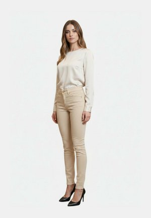 Donna con lunghi capelli castani che indossa una camicetta crema a maniche lunghe, pantaloni skinny beige e scarpe nere con tacchi alti, in piedi su uno sfondo bianco.