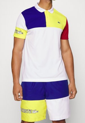 Mężczyzna ubrany w kolorowy polo i szorty w niebieskim, żółtym, białym i czerwonym kolorze, z logo Lacoste na piersi i lewym udzie.