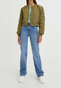 Woman wearing olive green bomber jacket, green and white striped shirt, light blue high-waist jeans, and white sneakers against a plain background.