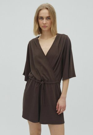 Nainen, jolla on olkapäille ulottuvat vaaleat hiukset, pukeutuneena tummanruskeaan lyhyeen kietaisumalliseen haalariin, jossa on vyötärönauha, seisoo vaalean yksivärisen taustan edessä.