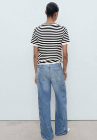 Randig svart och krämfärgad t-shirt med kort ärm som är parat med ljusblå vida jeans. Vistelsen är från baksidan mot en enkel bakgrund.