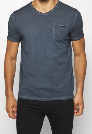 Mężczyzna ubrany w wyblakły, niebieski T-shirt z krótkim rękawem, z kieszenią po lewej stronie na piersi oraz czarne spodnie, na jednolitym tle.