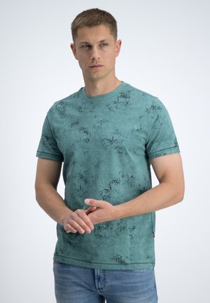 Ung mand iført turkis mønstret kortærmet skjorte og blå jeans, står mod en ensfarvet lys baggrund og kigger til siden.