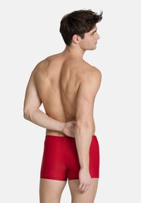 Rode zwemshorts met een gladde textuur, een aansluitend ontwerp en zonder zichtbare patronen. Model wordt van achteren getoond, waarbij de taille zichtbaar is.