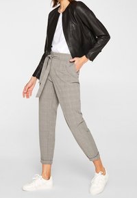 Chaqueta de cuero negra con cuello redondo, combinada con pantalones de cuadros grises ajustados y zapatillas blancas. Los pantalones tienen un cinturón de lazo.