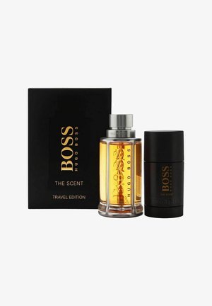 Hugo Boss The Scent Travel Edition sæt inkluderer en klar glasflaske med gylden væske, et sølvfarvet låg og en sort stiftdeodorant.