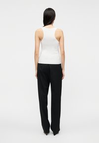 Γυναίκα με μακρ�ιά σκούρα μαλλιά, φορώντας λευκό αμάνικο ριπ τοπ και φαρδιά μαύρη παντελόνα, στέκεται με την πλάτη προς το φωτεινό ουδέτερο φόντο.