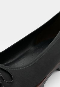 Black leather flat shoe with a rounded toe, textured upper, minimalist design, and a soft interior lining. Features a subtle lace detail.