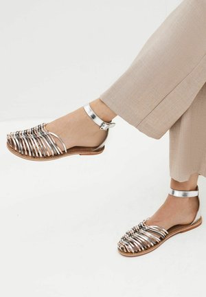 Metaliczne srebrne sandały z odkrytymi palcami, z wieloma poziomymi paskami i zapięciem na kostce. Gładka skóra syntetyczna, płaska podeszwa.