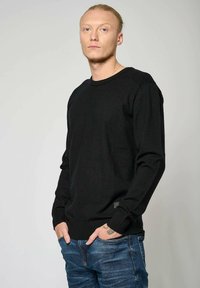 Pull noir à manches longues avec des détails côtes aux épaules et aux poignets, porté sur un jean bleu ; présente un petit logo brodé en bas.