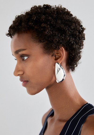 Srebrna ušna uhica u obliku kapljice s poliranim završetkom, prikazana na modelu s kratkom, kovrčavom kosom, ističući njezinu reflektirajuću površinu i konture.