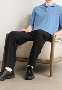 Polo bleu clair en maille, pantalon ample noir, chaussures noires à lacets, chaussettes blanches, assis sur une chaise beige recouverte de tissu avec des accoudoirs en bois.