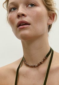 Smaragdovo zelený zamatový náhrdelník s obdĺžnikovými hnedými drahokamami, ozdobený striebornými detailmi; hladká textúra, tesný strih okolo krku.