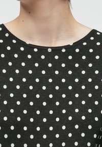 Svart stoffoverdel med rund hals og et mønster av hvite prikker, som gir et teksturert og visuelt tiltalende design.