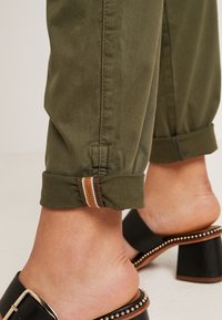 Pantalon vert olive roulé avec détail de languette rayée à la cheville, porté avec des sandales mules noires à talons ornées de clous dorés et d'une boucle.