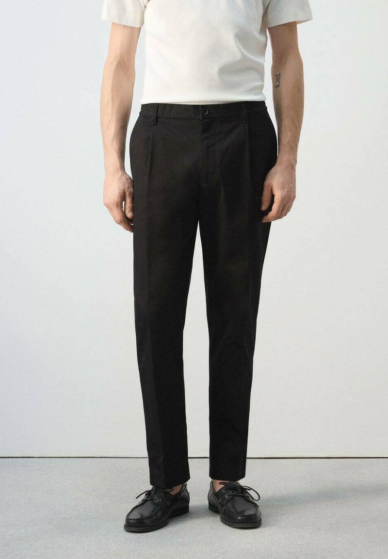 Mann trägt schwarze, maßgeschneiderte Hose, schwarze Schnürschuhe und ein weißes Kurzarmhemd, steht vor einem schlichten Hintergrund.