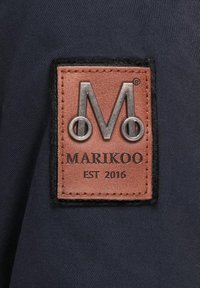 Brown leather label with embossed silver "M" and black "MARIKOO EST 2016" text, stitched onto dark fabric background.