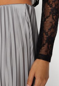 Šedá skládací sukně s hladkým povrchem, doplněná černým krajkovým topem s květinovými vzory a dlouhými rukávy. Ruka odpočívající na sukni.