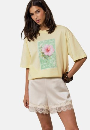 Kobieta w luźnym, bladożółtym T-shircie z różowym nadrukiem kwiatu hibiskusa oraz kremowych szortach z koronkową lamówką, pozująca z jedną ręką na biodrze.