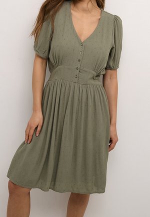 Femme portant une robe vert olive mi-longue avec des manches bouffantes, un décolleté en V, une boutonnière devant, une taille cintrée et un motif subtil à pois texturé.