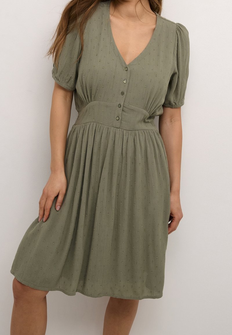 Femme portant une robe vert olive mi-longue avec des manches bouffantes, un décolleté en V, une boutonnière devant, une taille cintrée et un motif subtil à pois texturé.