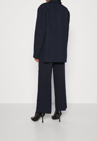 Donna che indossa un completo blu con righe verticali, pantaloni a gamba larga e scarpe con tacco alto nere, in piedi di fronte a uno sfondo semplice.