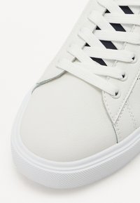 Valge nahast sneaker, millel on lamedad valged paelad, kummist talla ja tekstuuriga külgpaneelid, millel on geomeetriline disain.