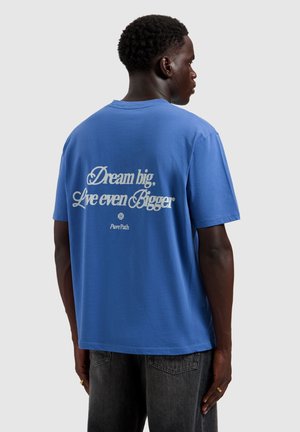 Mann mit dem Rücken zur Kamera, trägt ein blaues T-Shirt mit weißem Text „Dream big, Live even Bigger“ und schwarze Jeans vor einem schlichten hellen Hintergrund.