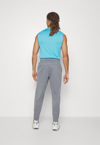 Man med dreadlocks som bär en ärmlös blå skjorta, grå joggingsbyxor, vita sneakers, står vänd mot en vit vägg.