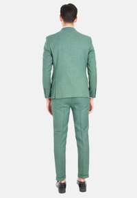 Completo verde testurizzato con design aderente, mostrato di spalle. Include pantaloni su misura e scarpe eleganti nere.