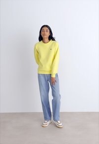 Sweatshirt amarelo com decote redondo e punhos canelados, combinado com jeans azul claro. O modelo usa ténis brancos e castanhos.