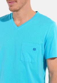 Jasnoniebieska koszulka typu V-neck wykonana z miękkiej bawełny, z kieszonką na lewej piersi z haftowanym logo. Gładka struktura, casualowy krój.