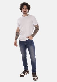 T-shirt bianco in cotone con scollatura rotonda e piccolo logo, abbinato a jeans in denim blu e sneakers marroni con dettagli bianchi.