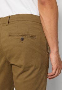 Chino maro cu o textură netedă, având un buzunar pe spate cu închidere prin buton și curea pentru centură. Design simplu și croială ajustată.