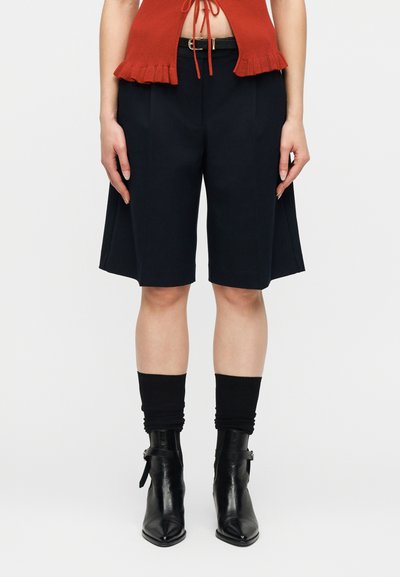 Femme portant un short noir cintré jusqu'aux genoux, une ceinture noire, des chaussettes noires larges, des bottines noires pointues et un haut court orange.