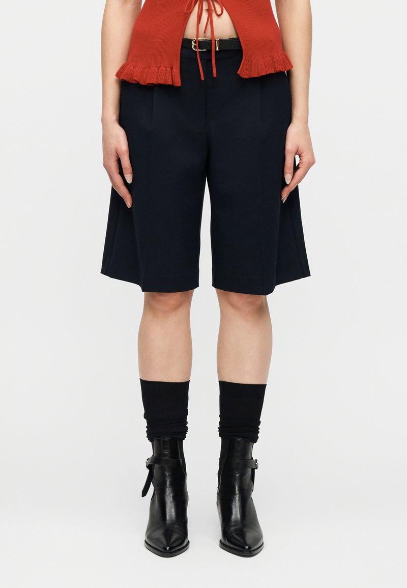 Γυναίκα που φοράει μαύρα, ραμμένα σορτς μέχρι το γόνατο, μαύρη ζώνη, μαύρες χαλαρές κάλτσες, μαύρα μυτερά μποτάκια μέχρι τον αστράγαλο και κομμένο πορτοκαλί τοπ.