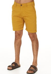 Gula bomullsshorts med uppvikta avslut, med knappstängning fram och sidofickor. Bärs med bruna sandaler på barfota.