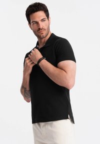 Muž s krátkými tmavými vlasy a strništěm nosí černé polo tričko, přizpůsobuje si límec. Má tetování a doplňuje svůj vzhled černým náramkem.
