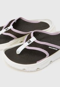 Ζευγάρι μαύρες και μωβ σαγιονάρες της μάρκας Salomon με υφασμένα πέλματα και λευκές παχιές σόλες σε απλό φόντο.