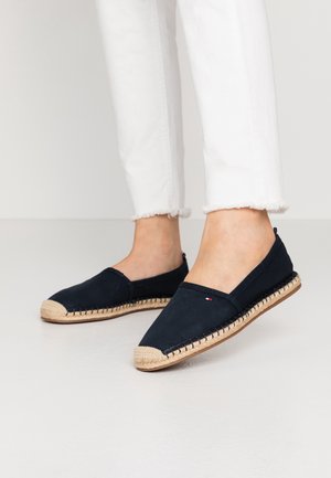 Personne portant des espadrilles noires à enfiler avec des semelles tissées beige et un pantalon blanc court avec des ourlets effilochés sur un sol clair.