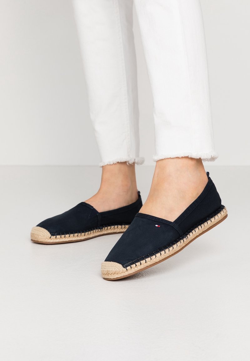 Personne portant des espadrilles noires à enfiler avec des semelles tissées beige et un pantalon blanc court avec des ourlets effilochés sur un sol clair.