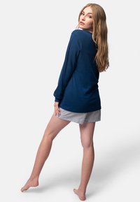 Maglia a maniche lunghe blu navy con texture a coste e spalle scese, abbinata a pantaloni corti a righe chiare, vista da dietro su uno sfondo neutro.