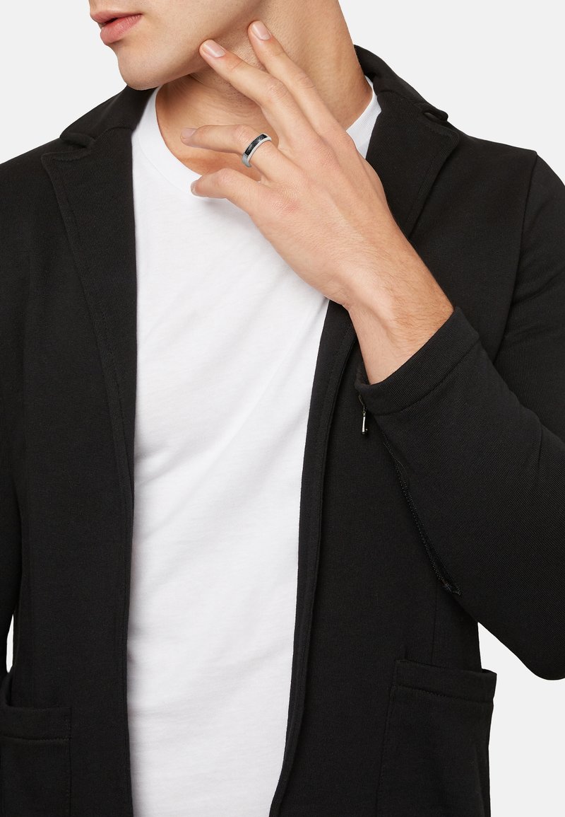 Blazer negro con una textura suave, con solapa muestrera, dos bolsillos frontales y un detalle de cremallera en la manga, sobre una camiseta blanca.