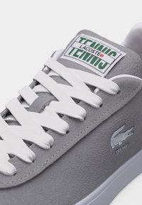 Sivá semišová teniska Lacoste s bielymi šnúrkami, štítkom „TRAINING TENNIS“ na jazyku a bielym logom krokodíla s nápisom „EST. 1933“ na boku.