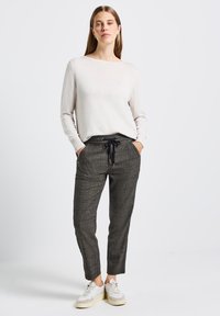 Vrouw staat met handen in de zakken, gekleed in een beige langarmtop, grijze geruite broek met trekkoord en witte sneakers op een witte achtergrond.