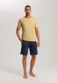 Gul bomulls T-shirt tillsammans med blårandiga shorts som har små vita och ljusblå motiv, modell stående barfota i en avslappnad pose.