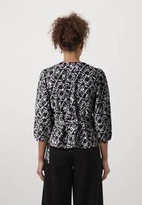Blouse à motifs noir et blanc avec des manches trois quarts, épaules froncées et ceinture à la taille, présentant un tissu lisse et texturé.