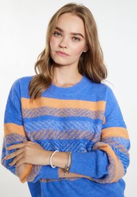Mėlynas ir oranžinis dryžuotas megztinis su apvaliu kaklu, laisva iškirpte ir tekstūruotu raštu. Modelis vilki apyrankes, matoma švelni medžiaga.