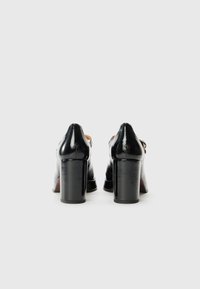 Escarpins en cuir verni noir avec un talon épais, texture lisse, bout rond et design minimaliste. Pas d'embellissements visibles.